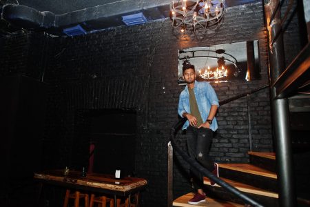 Portrait of handsome successful bearded south asian, young indian freelancer in blue jeans shirt standing in night club on stairs.